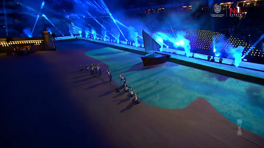 A dhow set piece on the projection-mapped pitch surrounded by performers and blue light