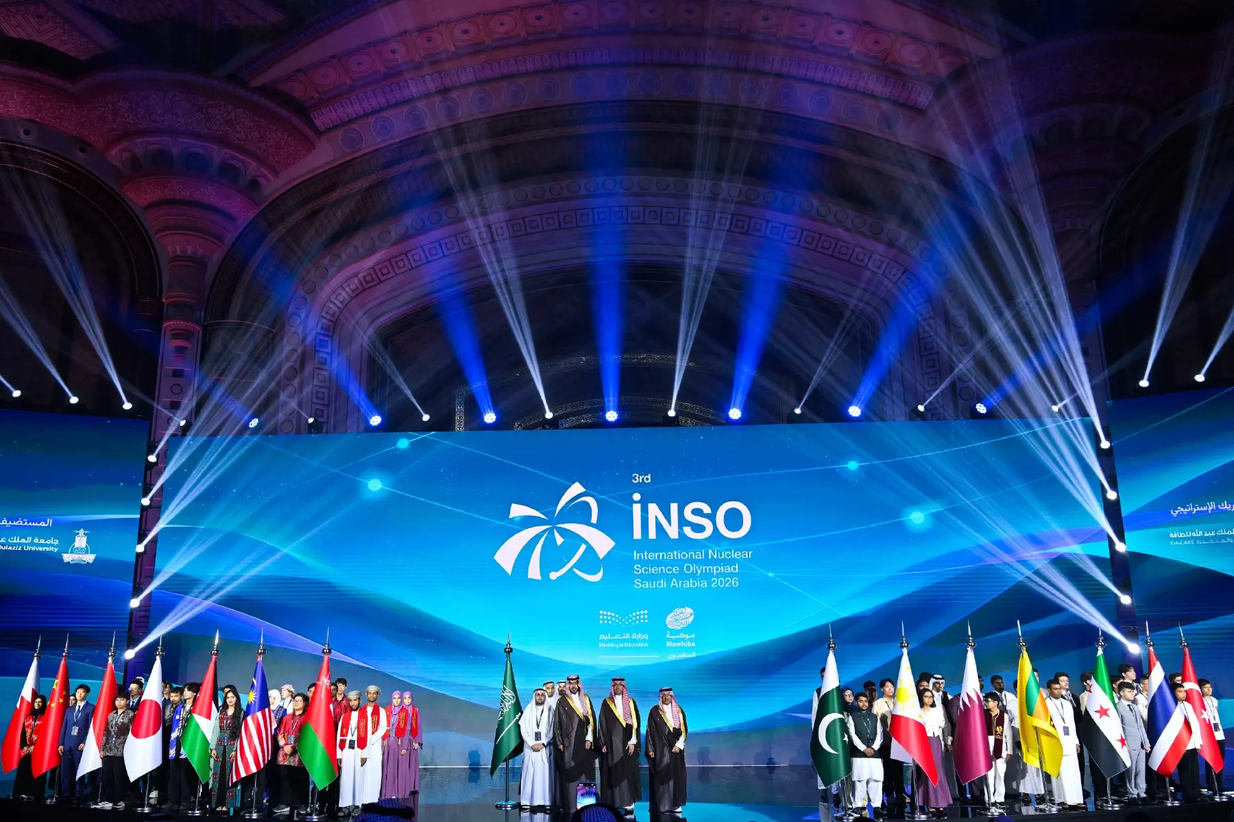 Blue beam fans across the domed ceiling of the King Faisal Conference Center above the INSO 2026 stage and flag bearers