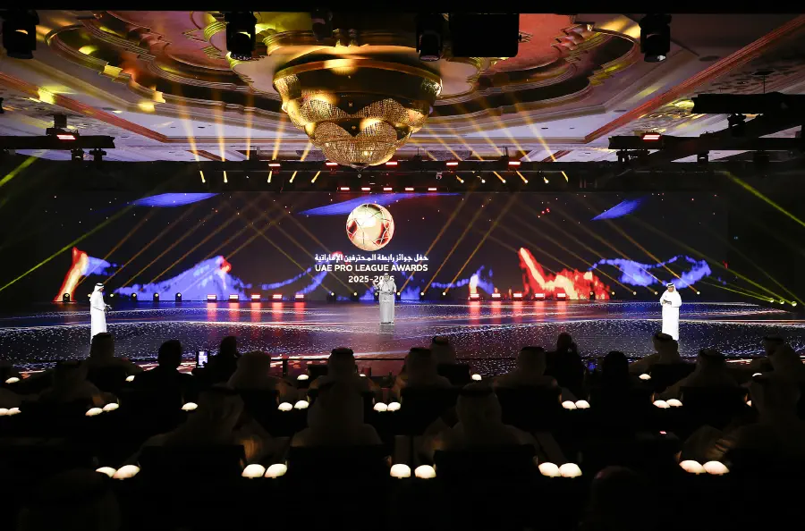 Wide view of the ballroom with tribunes around the LED floor, a presenter mid-stage and the audience in silhouette