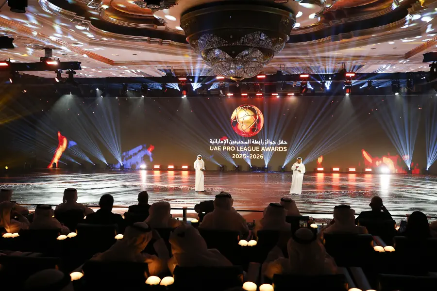 A presenter on the LED pitch under warm beams with the awards title on the main screen