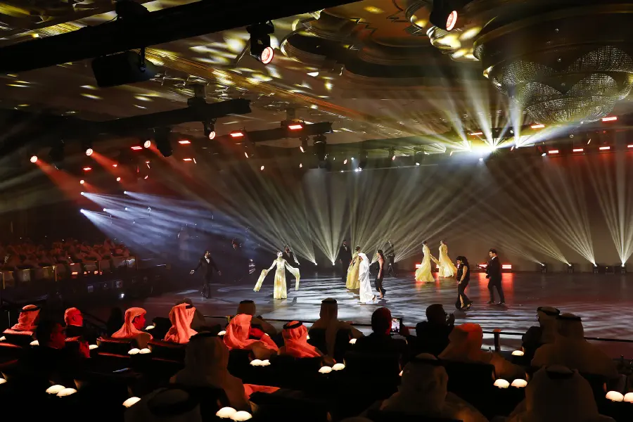 Performers on the LED pitch under a fan of warm beams, the audience lit in red